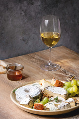 Fototapeta premium Cheese plate assortment of french cheese served with honey, walnuts, bread and grapes on ceramic plate and glass of white wine over wooden table with grey wall as background.