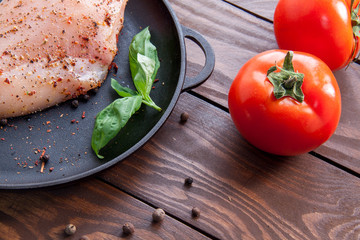 ripe red tomatoes on a wooden table next to a frying pan on which a fresh raw chicken fillet is sprinkled with herbs and spices