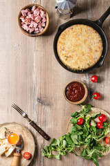 Spanish potato omelette tortilla with bacon served in cast-iron pan with sauce and ingredients above over wooden background. Top view, space.