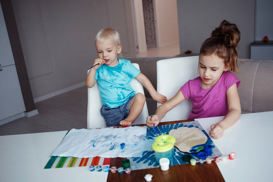 Two happy children blond boy and girl drawing paints and brushes and sitting at table at home.