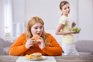 Being an outcast. Miserable stout woman eating a sandwich and her slim friend smirking