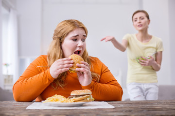 Mockery. Unhappy stout woman eating a sandwich and her slim friend criticizing her