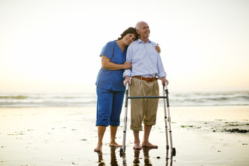 Senior man being comforted by a female nurse while at the beach with his walking aid.