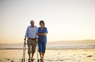 Senior man walking with a walking aid next to a female nurse on the beach.
