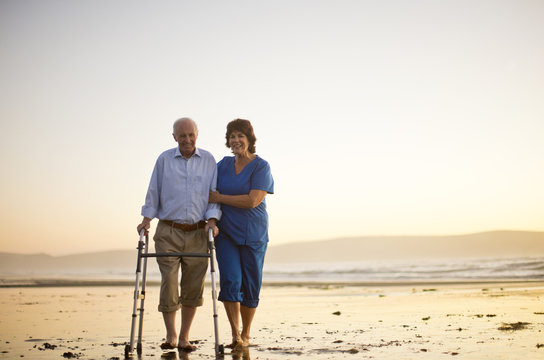 Senior man walking with a walking aid next to a female nurse on the beach.