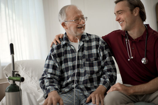 Cheerful elderly man with a nasal tube speaking with a male nurse at his hospital bed.