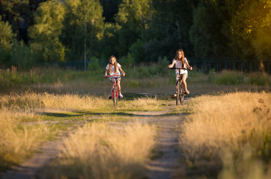 Two smiling teenage girls riding bicycles in the field at sunset