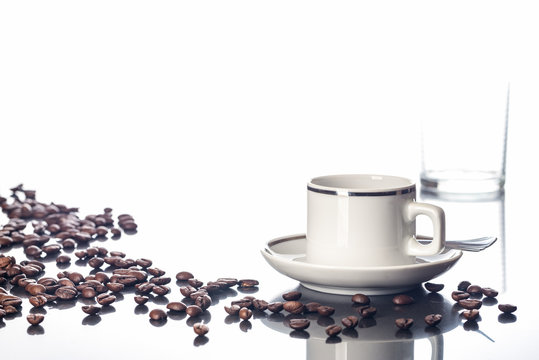 Espressotasse, Kaffeebohnen und Wasserglas auf spiegelnder Tischplatte vor weißem Hintergrund
