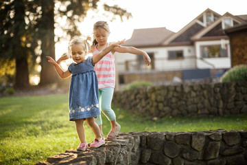 Two sisters playfully balancing on a stone wall.