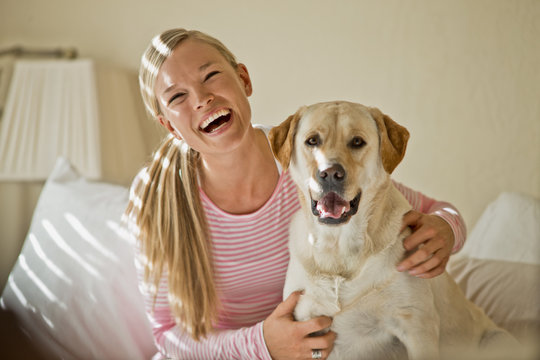 Happy young woman laughs as she hugs her dog in her sunlit bedroom.