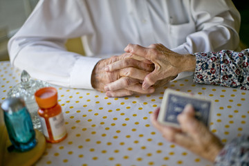 Elderly wife gently places her hand atop her husband's hands as they play cards together at the kitchen table.
