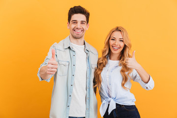 Portrait of cheerful people man and woman in basic clothing smiling and showing thumbs up at camera, isolated over yellow background