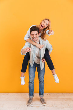 Full length photo of smiling couple having fun and pointing fingers at you while man piggybacking happy woman, isolated over yellow background