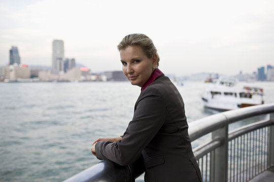 Portrait of a mid-adult businesswoman standing at a railing overlooking a city harbor.