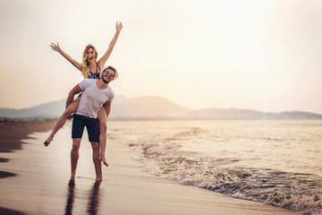 Happy couple in love on beach summer vacations. Joyful girl piggybacking on young boyfriend having...
