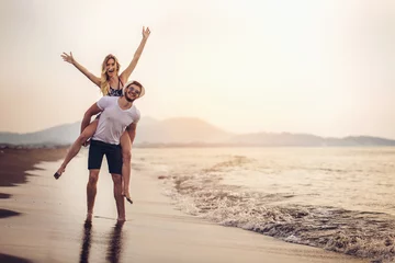 Fotobehang Happy couple in love on beach summer vacations. Joyful girl piggybacking on young boyfriend having fun. © Mediteraneo