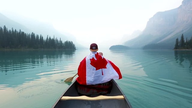 A white mal with a cap wrapped in the Canadian flag paddles over a lake in a kajak