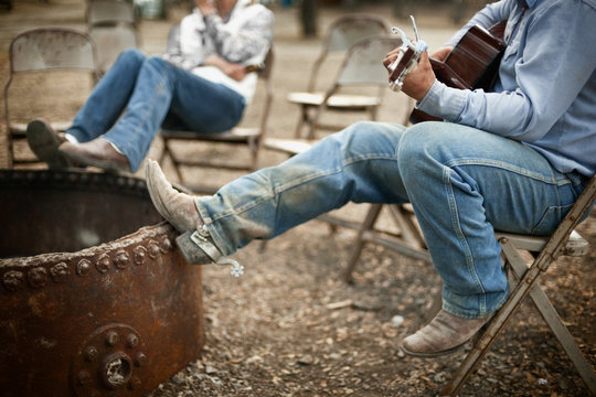 Man plays guitar as he and a male friend relax resting their spurred cowboy boots on the edge of an empty outdoor metal firepit.