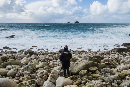 A woman standing on the rocks looking out over the Cornish coastline, and breaking waves. 