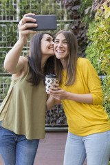 two beautiful women taking a selfie with mobile phone. One is holding a cup of coffee and kissing her friend. They are laughing. Outdoors lifestyle and friendship concept
