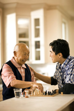 Senior man makes a chess move and his younger opponent puts an affectionate hand on his shoulder as they smile and talk over a chess set while they sit at the end of a kitchen counter.