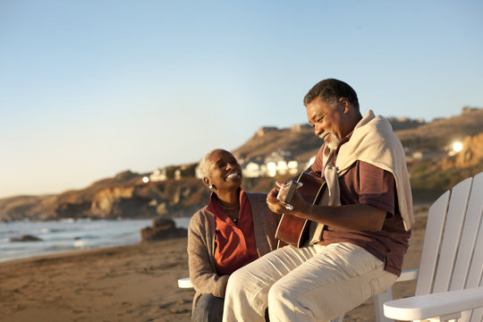 Smiling senior woman listening to her husband play an acoustic guitar on a beach.
