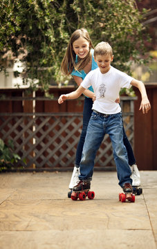 Girl holding onto her younger brother's waist as they try to roller skate.