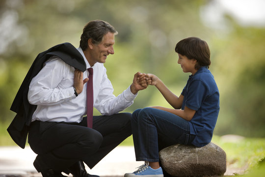 Businessman father crouches down to knock fists together (daps) with his young son who sits on a rock in a park.