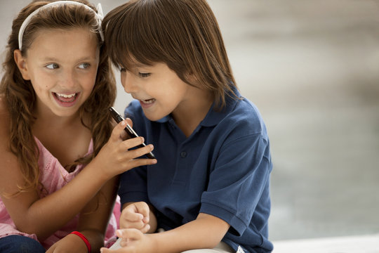 A young boy and girl smile mischievously as they look at a cellphone.
