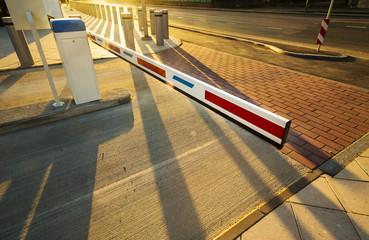 Close up of boom barrier gate entrance illuminated by warm sunset light. Perspective view with long shadows