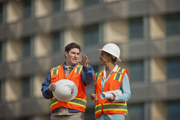 Two engineers in high visibility vests and hardhats discuss building plans on a construction site.