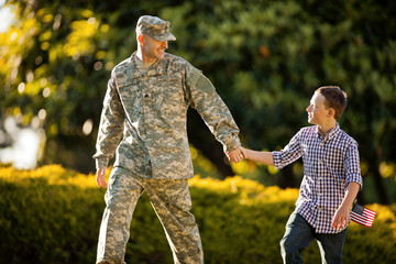 Army soldier holding hands with his young son in their back yard.