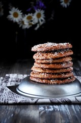 On a wooden table a stack of curd cookies with coconut chips. Decorated with flowers. On a metal plate. Dark background