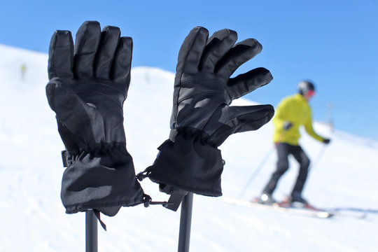 Detail on black ski gloves on ski poles with blurred skier in background on bright sunny winter day.