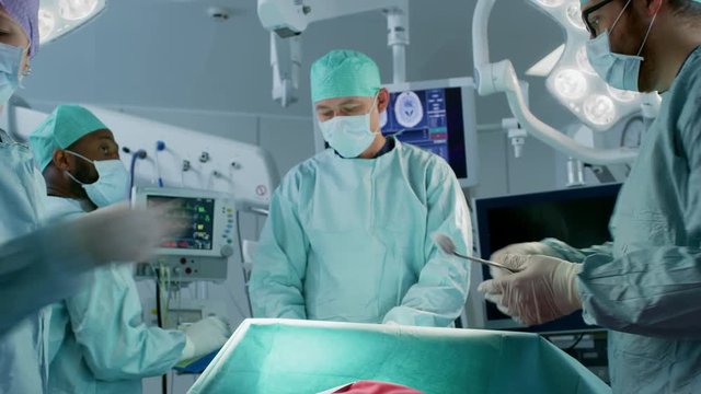 Diverse Team of Professional Surgeons Performing Invasive Surgery on a Patient in the Hospital Operating Room. Nurse Passes Instrument to a Chief Surgeon, Anesthesiologist Monitors Vitals. 