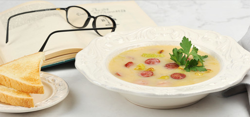 Potato soup with sausages in a deep dish. Next to the plate with toast of white bread. In the frame is a book and glasses. Close-up. Light background.