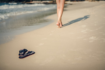 The girl walks barefoot along the beach. Flip-flops on the sand.