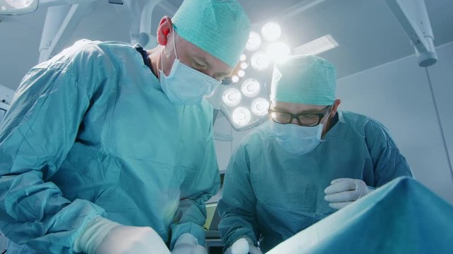 Low Angle Shot In Operating Room of Two Surgeons During the Surgery Procedure Bending Over Patient with Instruments. Professional Doctors in Modern Hospital