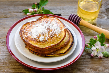 Sweet homemade pancakes with powdered sugar on a plate decorated with flowers of an apple tree on a wooden background.