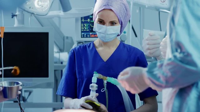 In the Hospital Operating Room Anesthesiologist Applies Anesthesia Mask to a Patient, Assistants Disinfects with Iodine place of Incision, Surgeons Wait to Start Surgery.