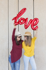 two beautiful young women having fun outdoors with a red balloon with a love word shape. Casual clothing. They are wearing hats and modern sunglasses. LIfestyle outdoors