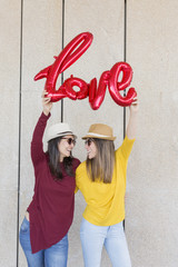two beautiful young women having fun outdoors with a red balloon with a love word shape. Casual clothing. They are wearing hats and modern sunglasses. LIfestyle outdoors