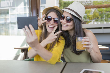 two beautiful women having breakfast in a restaurant and taking a selfie with mobile phone. They are laughing. Indoors lifestyle and friendship concept