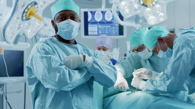 Professional Surgeon Standing in Surgical Mask Arms Crossed. In the Background Modern Hospital Operating Room with Surgery in Progress.