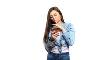 Fototapeta premium Portrait of a young brunette woman looking at the smartphone in her hands with hesitation and wistfulness, thinking against white background, isolated. Lifestyle, people and technology concept