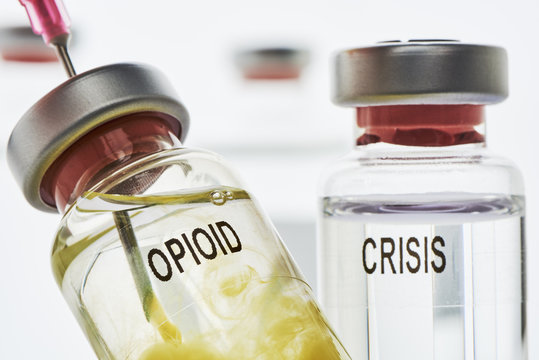 Opioid Krise - In eine Opioid Ampulle die klare flüssigkeit enthält, wird mit einer Nadel eine gelbeflüssigkeit injeziert. Im hintergrund ist ein Ampulle mit der Aufschrift Krise