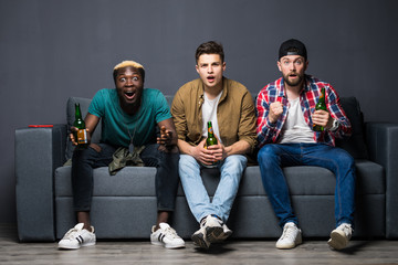 Fototapeta premium Group of exited men sitting together on couch, watching television holding beers, screaming