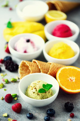 Top view red, pink, yellow, green, white ice cream balls in bowls, waffle cones, berries, orange, mango, pistachio, grey concrete background. Colorful collection, flat lay, summer concept
