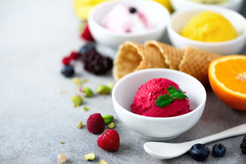 Top view red, pink, yellow, green, white ice cream balls in bowls, waffle cones, berries, orange, mango, pistachio, grey concrete background. Colorful collection, flat lay, summer concept