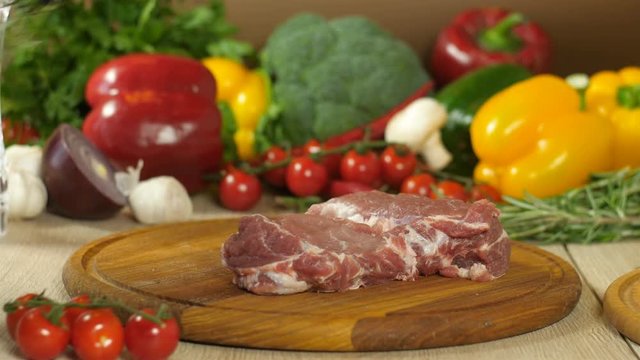 The chef in black gloves lays a steak on a round wooden plank, from above lays a sprig of rosemary, a clove of garlic and sprinkles with a mixture of peppers, in the background vegetables, the